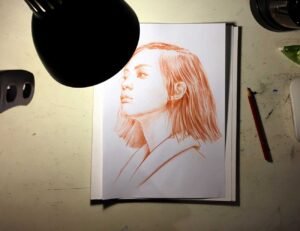 Work in progress view of “Terracotta Light” – red pastel female portrait drawing under studio light